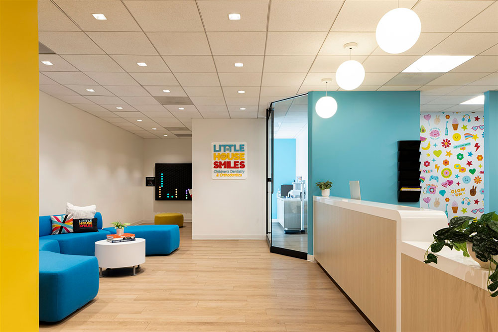 Bright pediatric dental lobby with blue seating, reception desk, glass office, and colorful wall graphics