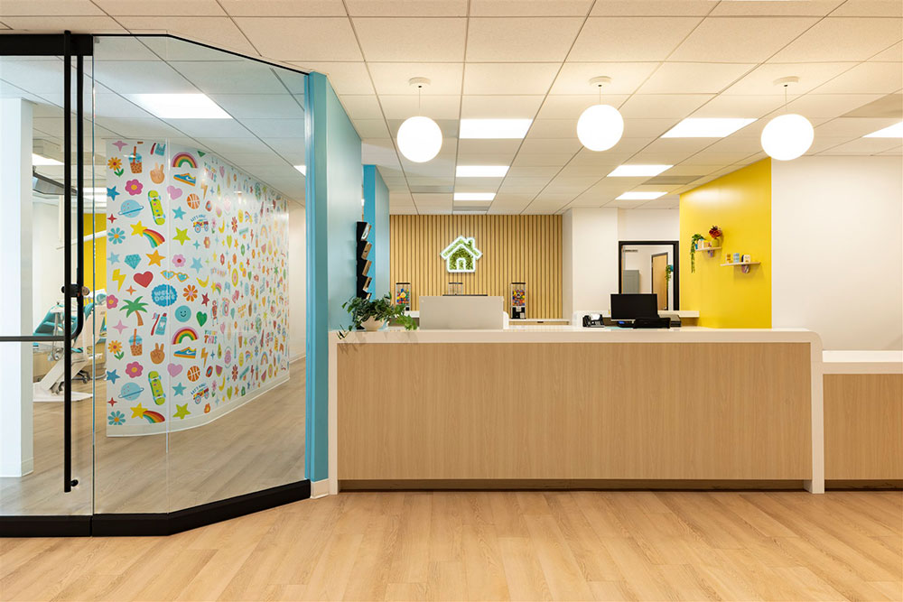 Pediatric dental reception desk with wood finishes, yellow accent wall, glass partition, and colorful hallway mural