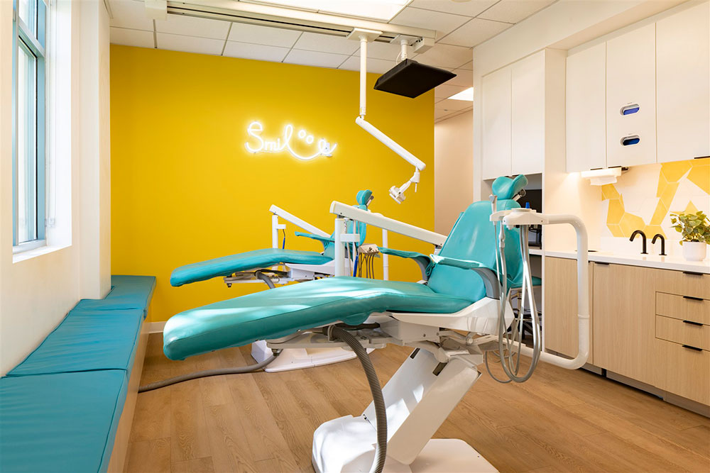 Modern pediatric dental exam room with dual teal chairs, yellow accent wall, neon smile sign, and built-in cabinetry