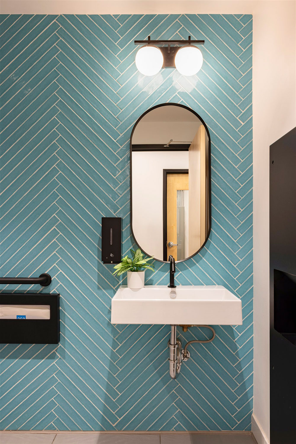 Modern restroom with wall-mounted sink, oval mirror, black fixtures, and blue herringbone tile accent wall