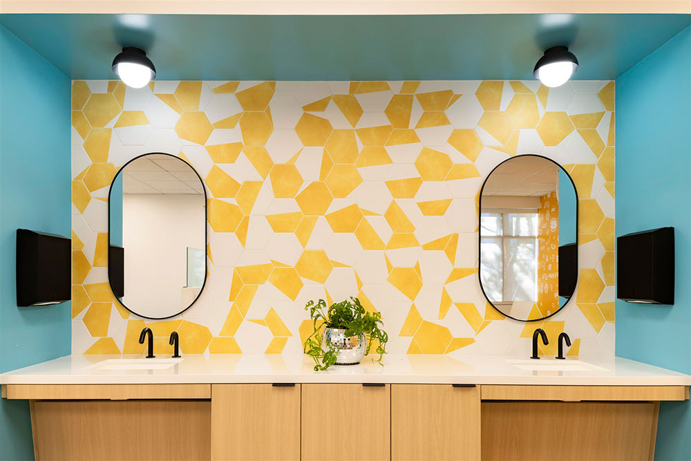 Modern restroom with double sinks, oval mirrors, wood cabinetry, and yellow geometric backsplash in a bright office space