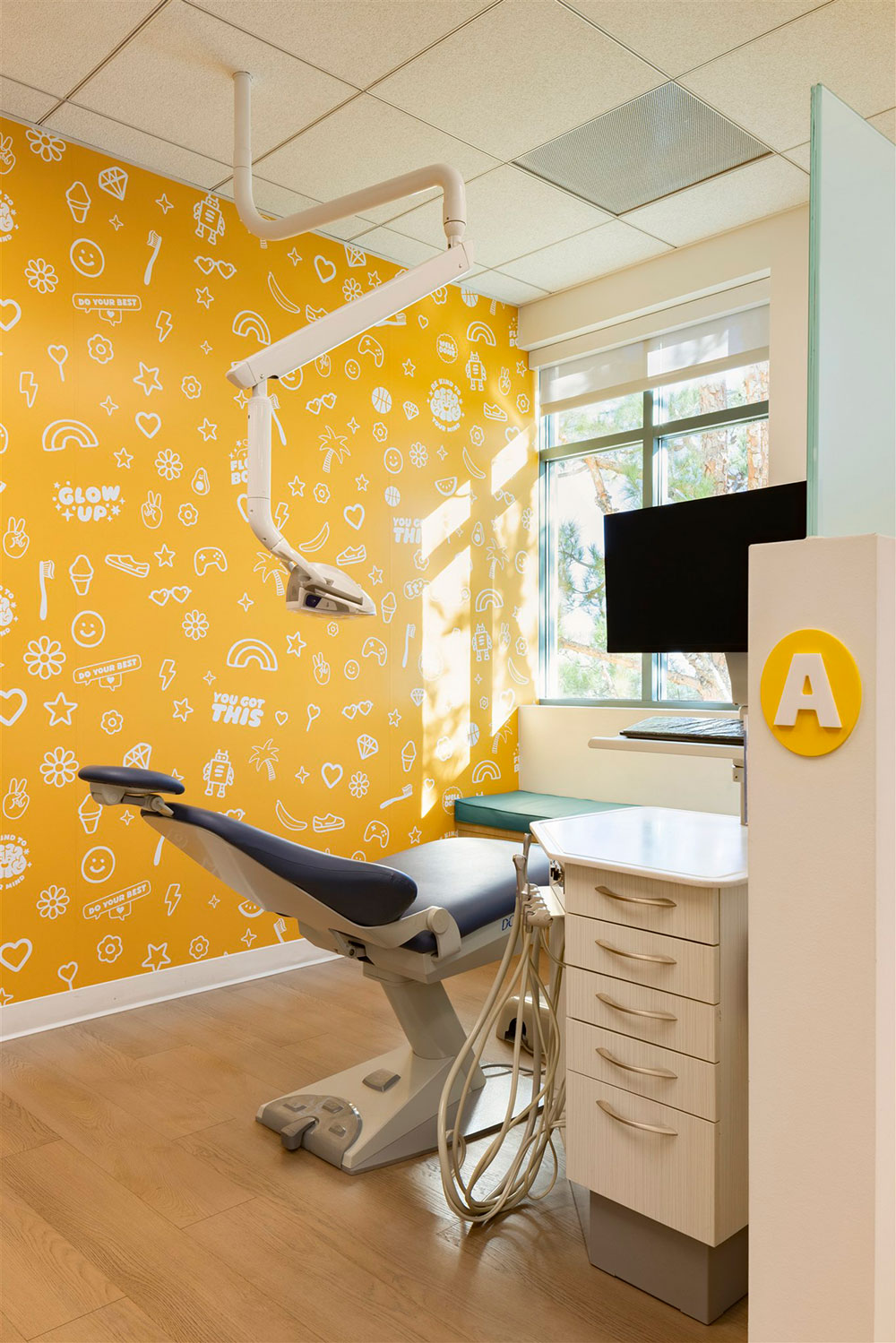 Colorful pediatric dental exam room with patterned yellow wall, dental chair, overhead light, and modern workstation