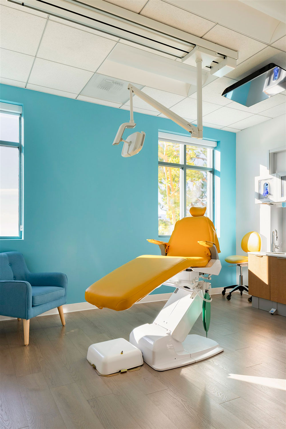 Bright dental exam room with yellow chair, blue walls, overhead light, and modern equipment by window