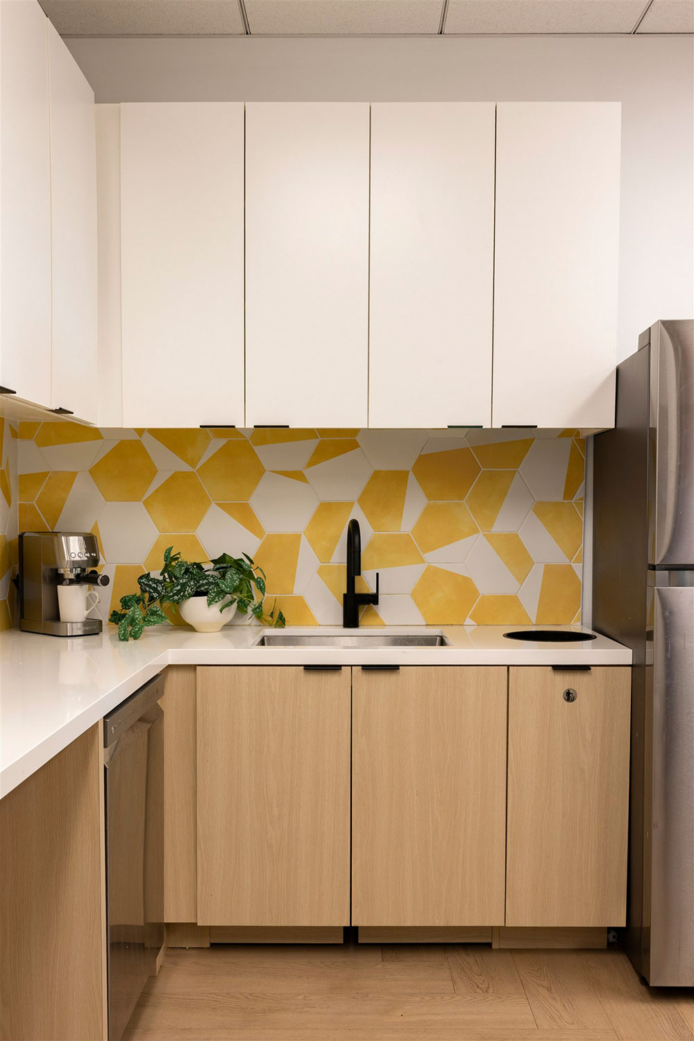 Modern office kitchenette with white cabinets, wood base, yellow geometric backsplash, sink, coffee machine, and fridge